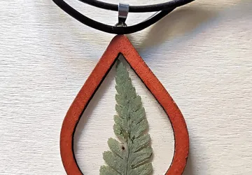 Colla de bisel de madera de tonos rojizos con hoja de helecho encapsulada en bio resina. Cordón de cuero encerado negro.